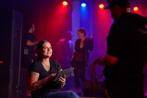 Eine Frau mit einem Tablet in einem dunklen Raum mit Scheinwerfern, im Hintergrund eine Band